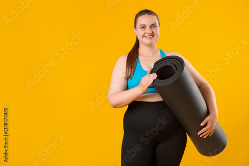 Cuadro en lienzo Happy overweight woman with fitness mat on yellow background, space for text
