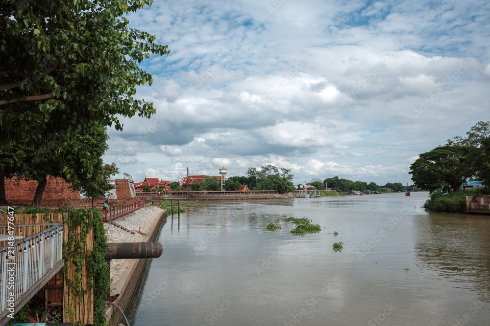 Fototapeta premium A scenic exterior view of Phra Nakhon Fortress located between the Chao Phraya River and the Pasak River in Ayutthaya, Thailand, featuring historic brick walls, a wooden waterfront structure, trees, a
