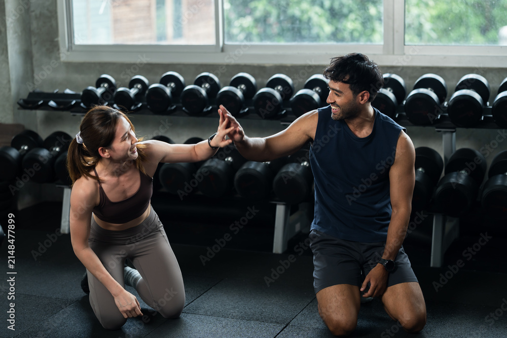 Fototapeta premium Young Asian couple celebrates partner workout with high five on modern gym floor, man and woman sharing motivation, teamwork, encouragement, healthy relationship, bodyweight strength lifestyle.