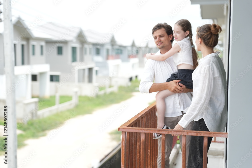 Obraz premium Young parents holding their child enjoy the view of the growing suburb from their balcony standing on the terrace of a newly built suburban home. Happy family lifestyle and wellbeing concept