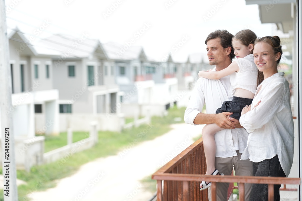 Obraz premium Happy young family with little daughter look out over the rows of new white houses from the railing standing on the terrace of a newly built suburban home, shallow depth of field