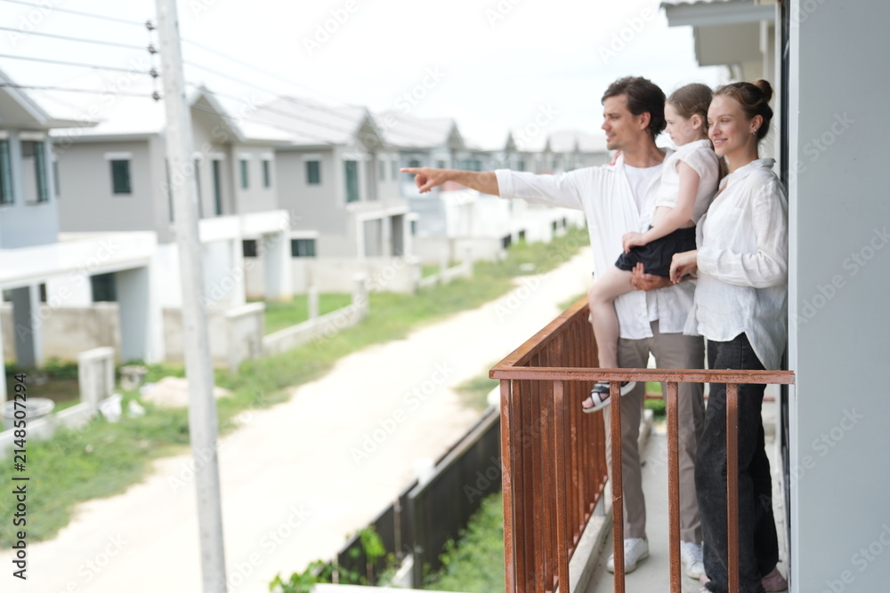 Obraz premium Young parents holding their child look out over the rows of new white houses from the railing on the upper balcony of their new home above the neighborhood in warm natural light