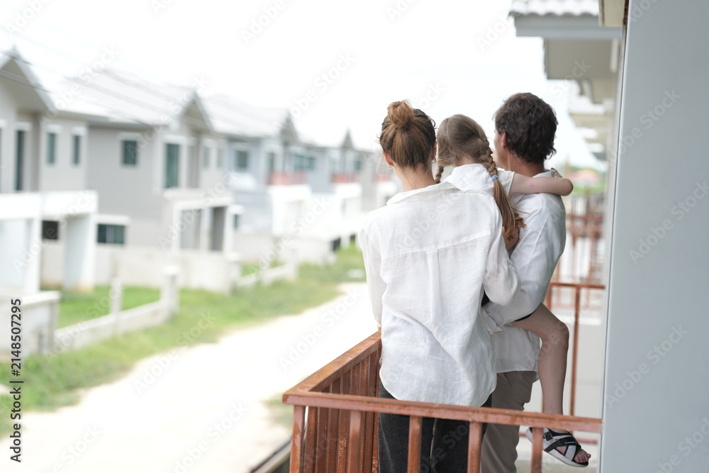 Obraz premium Happy young family with little daughter stand at the wooden railing admiring the neighborhood view standing on the terrace of a newly built suburban home, shallow depth of field