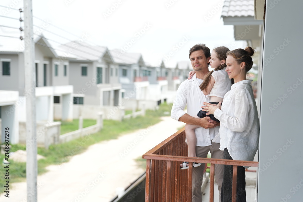 Obraz premium Young parents holding their child stand at the wooden railing admiring the neighborhood view on the upper balcony of their new home above the neighborhood on a bright sunny day