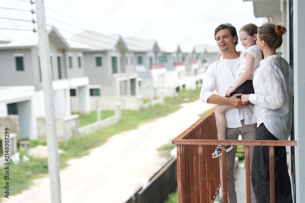 Obraz premium Caucasian family of three embracing stand at the wooden railing admiring the neighborhood view on the upper balcony of their new home above the neighborhood on a bright sunny day