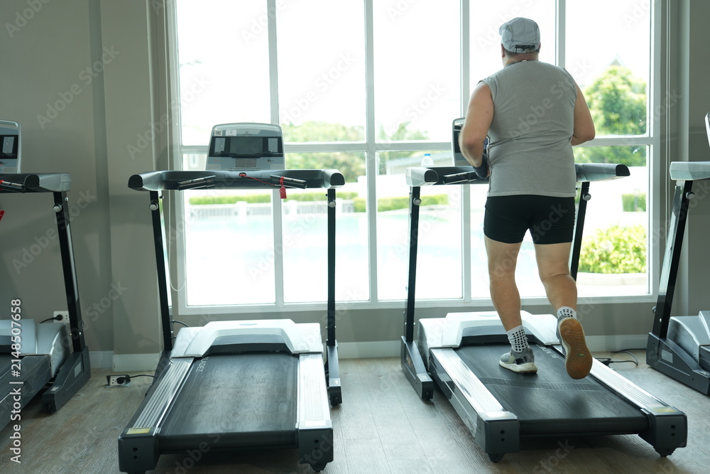 Fototapeta premium Overweight middle aged man in gray cap and sleeveless shirt raises his arms in victory while standing on the treadmill at the start of his new healthy lifestyle in the gym