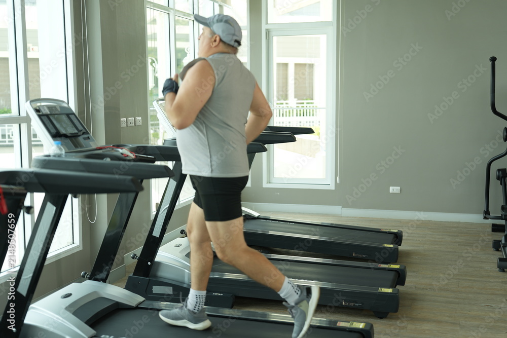 Fototapeta premium Heavyset mature man determined to lose weight does his cardio walk on the treadmill machine in a bright modern gym with large windows. Funny weight loss and healthy lifestyle concept