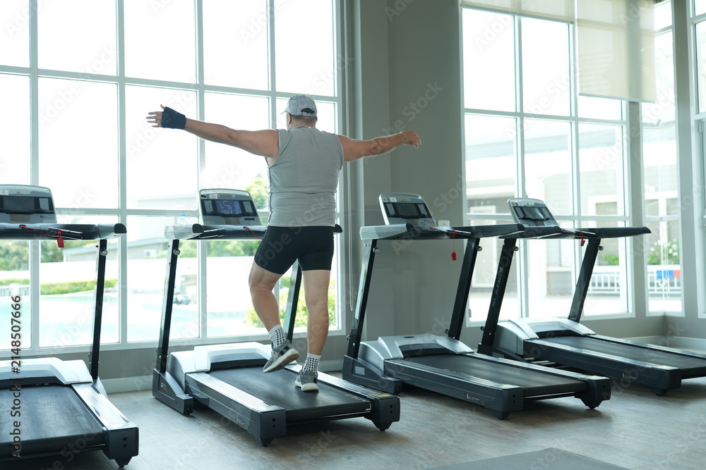 Fototapeta premium Funny fat senior man in sportswear balances playfully on the treadmill with arms spread wide during his weight loss workout at the fitness club in warm natural light