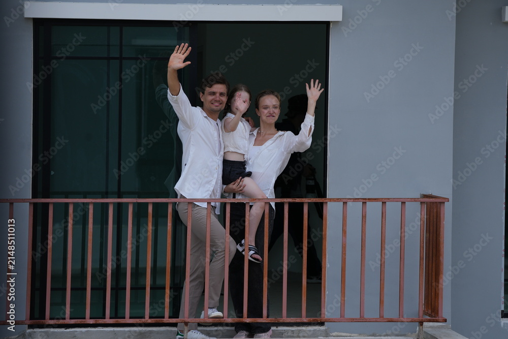 Obraz premium Caucasian family of three embracing laugh and wave from the new balcony railing standing on the terrace of a newly built suburban home. Healthy active lifestyle of a modern family