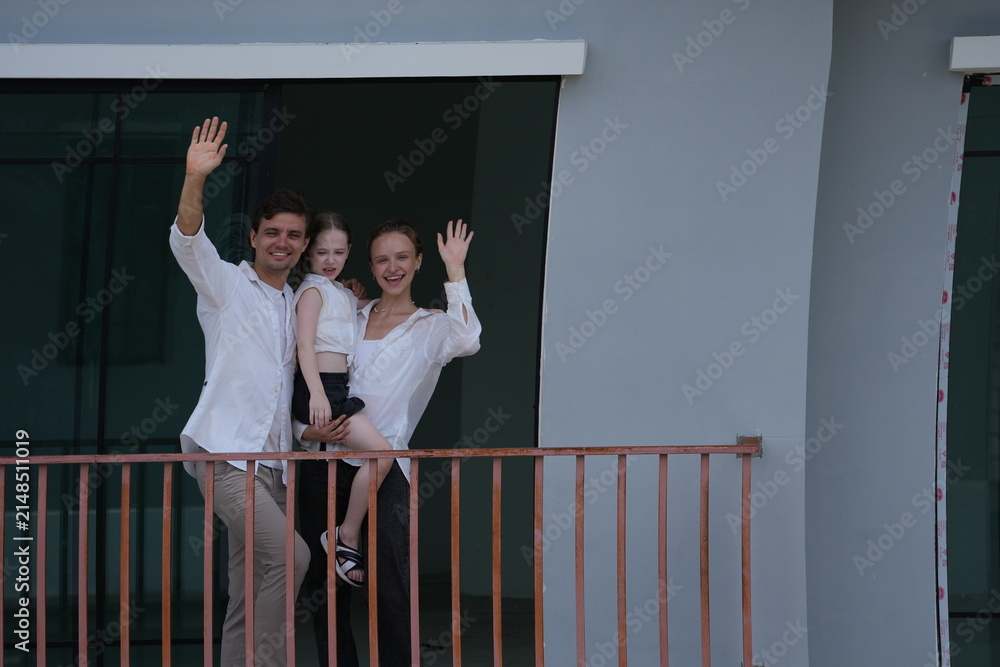 Fototapeta premium Caucasian family of three embracing wave happily from their balcony, the girl riding piggyback on the balcony of their new house overlooking the residential estate, authentic everyday scene