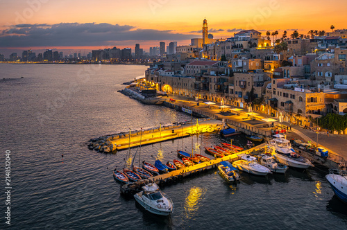 Aerial view of Jaffa Port at sunset with colorful fishing boats at the pier, historic stone buildings, and the city skyline in the background, Tel Aviv-Jaffa, Tel Aviv District, Israel.