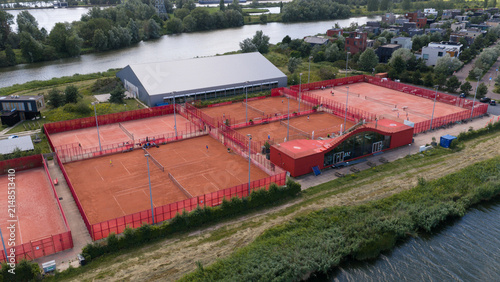 Aerial view of Tennisclub IJburg with its clay tennis courts, red fencing, and modern clubhouse situated along a canal in Amsterdam, Noord-Holland, Netherlands.
