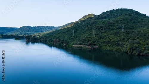 Aerial view of a wide river flowing past lush green hills with power transmission towers in Ghana.