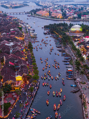 Aerial view of the Thu Bon River featuring numerous small wooden boats, illuminated historic buildings, and bustling waterfront streets at dusk in Hoi An, Vietnam.