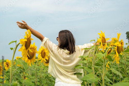 Young woman with shoulder length brown hair stands in sunflower field with arms raised, facing away under pale blue sky, surrounded by tall yellow blooms and green foliage in bright summer light