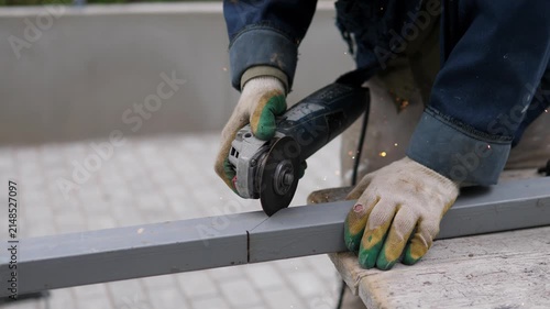 Close-up shows gloved worker pressing handheld grinder onto steel beam with bright sparks, dark workwear, worn wooden support, soft natural light, and blurred background in practical workshop