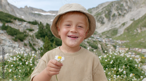 This outdoor shot shows a smiling young boy in a bucket hat holding a small daisy, set against a backdrop of alpine meadows dotted with wildflowers and distant rocky mountains.

