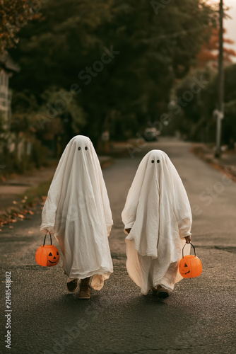 Two Children in Ghost Costumes Walking with Pumpkin Buckets on Residential Street at Dusk Creating a Cozy Halloween Atmosphere. Copy Space, vertical 2:3