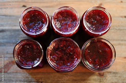 Six open glass jars filled with homemade red berry jam stand on a rustic wooden table