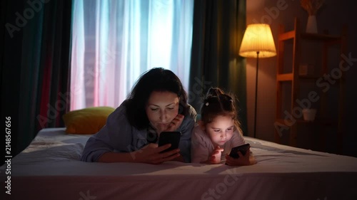 mother and daughter are using smartphones while lying on the bed in the evening.
