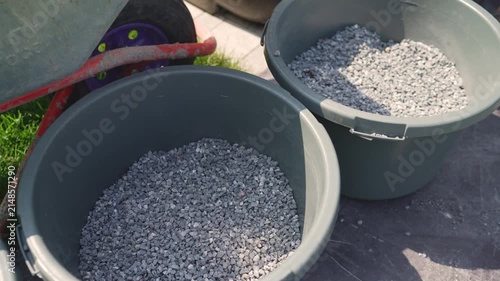Closeup of gray gravel inside dark plastic buckets on paved ground. Bright daylight highlights texture shadows. Green grass red wheelbarrow in background