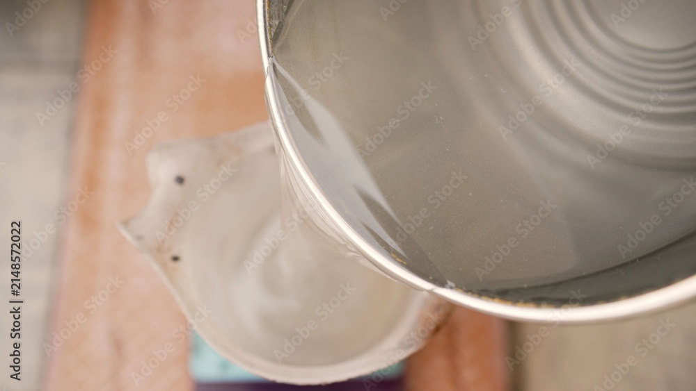 Obraz premium Overhead view of creamy white liquid flowing from metal bucket into ceramic vessel. Soft natural lighting, smooth texture, warm wooden background