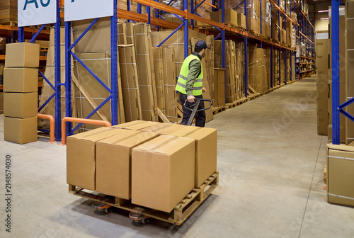 Warehouse worker moves pallet cartons on trolley. Organized storage logistics and shipping in a large center with high shelves and racking. Streamlined warehouse logistics and fast delivery.