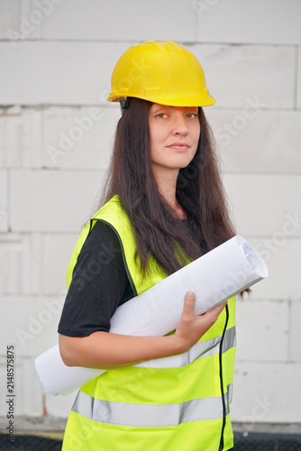 Young female construction worker in hi vis vest, helmet and gloves, carrying cable roll on her shoulder, bare new house wall near