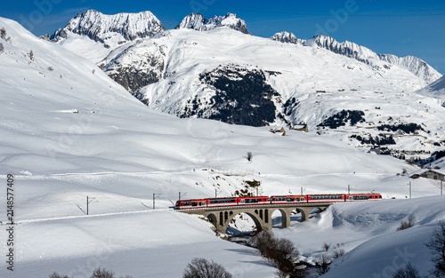 Aerial view of the Glacier Express train crossing a stone arched viaduct in a snowy mountain landscape in Bergun Filisur, Graubunden, Switzerland.