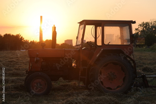 Konstfotografi Old red tractor in a mown hayfield with a power plant behind at sunset