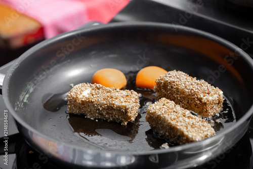 Three pieces of sesame-coated food cooking in a frying pan with two raw egg yolks beside them on a stovetop in a kitchen setting