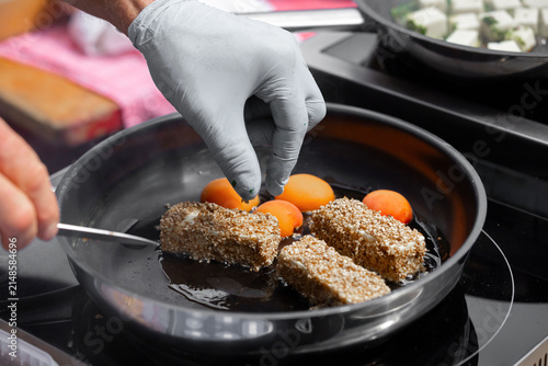 Chef in gray gloves carefully places sesame-coated food items and orange cherry tomatoes in a frying pan on a modern stovetop during food preparation