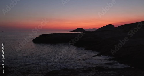 Soft orange and red light up the pre dawn morning as the sun rises over the island of Milos, Greece. Aegean sea water gently flows to the shore of Mandrakia looking toward Sarakiniko beach. Quiet 