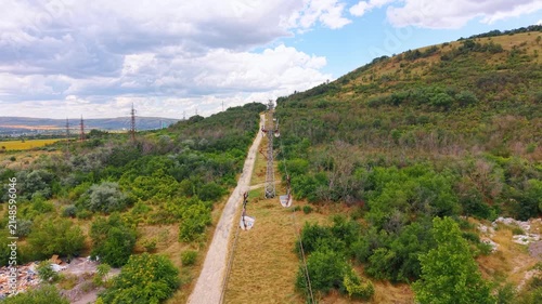 Cableway pylons extending up grassy hillside. Wide aerial view shows industrial steel towers and cable lines above countryside path.