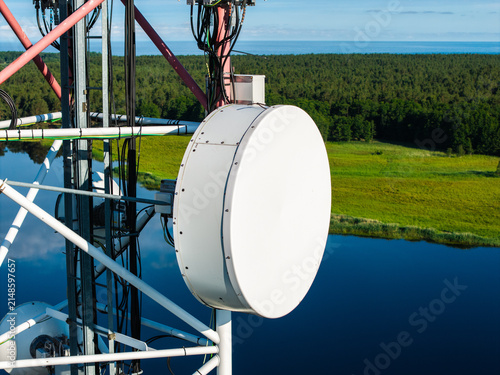 Aerial view of a large white parabolic antenna mounted on a telecommunications tower overlooking the Bullupe River with lush green banks and forest in Riga, Latvia.