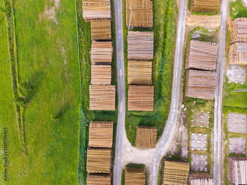 Aerial view of neatly stacked timber logs in a forestry area, surrounded by green grass and narrow access roads in Latvia.