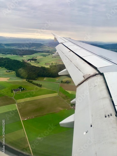 Airplane wing flying over green landscape fields view from window.