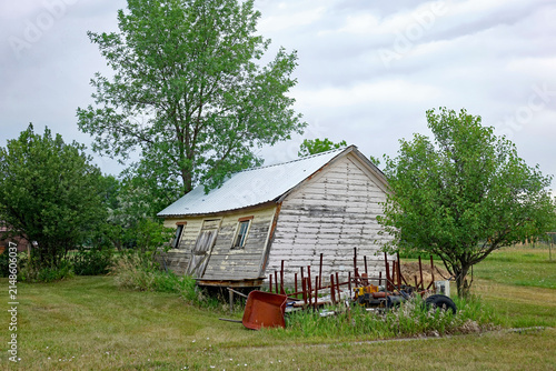Leaning Abandoned White Shed With Rusty Wheelbarrow and Tools in Quiet Rural Yard