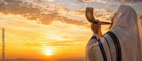 Religious Jewish man wrapped in a tallit blowing the shofar horn against a dramatic sunset sky. Background concept for Rosh Hashanah (Jewish New Year), Yom Kippur and High Holy Days.