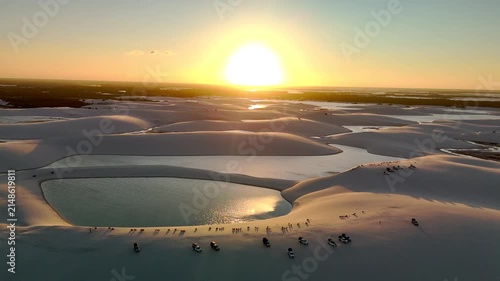 Aerial view of Parque Nacional dos Lencois Maranhenses featuring vast white sand dunes, crystal clear lagoons, and off-road vehicles at sunset in MA, Brazil.