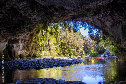 View of the Oparara Arch cave entrance with a calm river flowing through the rocky riverbed and lush native forest visible beyond in Karamea, West Coast Region, New Zealand.