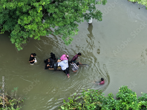 Banshkhali Upazila, Chittagong Division, Bangladesh - 10 July 2026: Aerial view of people wading through floodwaters, pushing a makeshift raft through murky water near lush trees.