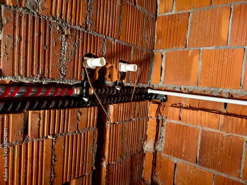 Water and heating pipes installation with red and blue insulation on a red hollow brick wall. Rough plumbing work on an under-construction building site.