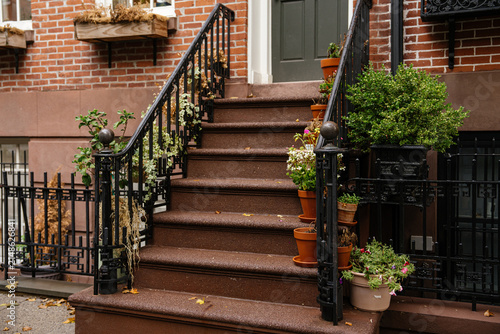 Close up view of brownstone stairs with black iron railings and multiple potted plants in a New York City neighborhood. Brick facade and decorative details highlight classic urban residential