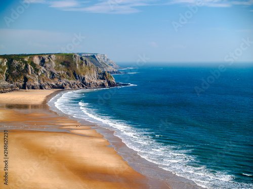 Aerial view over Three Cliffs Bay, golden sand beach curving beneath limestone headland, turquoise sea, Gower Peninsula, Wales.