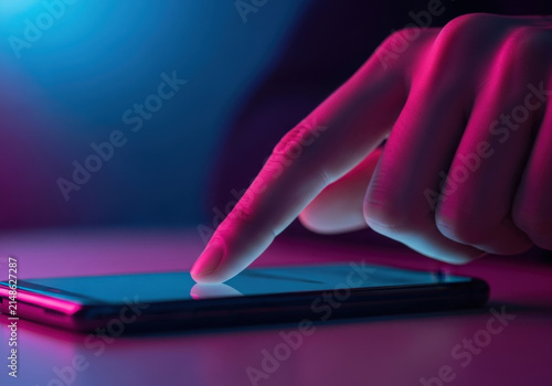Finger touching smartphone screen with pink and blue neon lighting in dark room atmosphere showing mobile application usage digital technology.