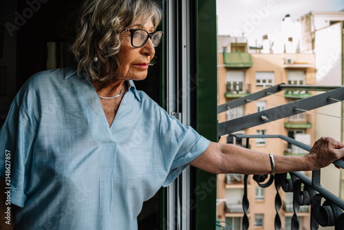 Elderly woman looking out city balcony feeling lonely and thoughtful