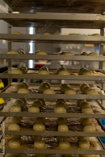 multiple rows of smooth proofed dough balls arranged on metal baking trays within tall bakery racks, lit by warm dim kitchen light with soft focus equipment behind