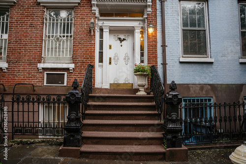 A traditional New York brownstone entrance featuring stone steps, ornate black iron railings, and a white decorative door. Wet sidewalk and brick facade highlight historic urban residential