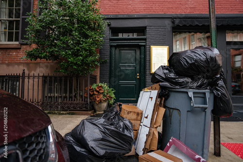 Black garbage bags and cardboard boxes stacked beside a trash bin on a New York City sidewalk. Urban street scene shows residential building entrance and everyday city life.